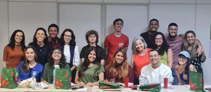 Na foto, estagiários da SEAD agrupados ao centro, todos sorriem e formam uma fila de alunos sentados por trás de uma mesa e uma fila de alunos de pé abraçados. O grupo é diverso com pessoas do sexo feminino e masculino e de várias cores e tipos de cabelo e roupas